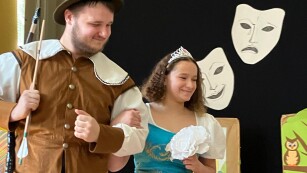 Na pierwszym planie widać aktorów, dziewczyna wciela się w rolę królewny z bajki o „Królewnie Śnieżce”, chłopiec w myśliwego. Dziewczyna ma piękną suknię z bufiastymi rękawkami,, w ręku trzyma kwiaty-róże. Aktor grający myśliwego ma łuk i strzałę, na głowie kapelusz myśliwski i kubrak. W tle widać maski teatralne stanowiące część dekoracji.