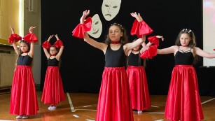 Na scenie stoją tancerki prezentujące taniec hiszpański. Jest ich 5 i są ubrane jednakowo: w długie, rozkloszowane spódnice i koszulki bez rękawków, mają uniesione ręce do góry.