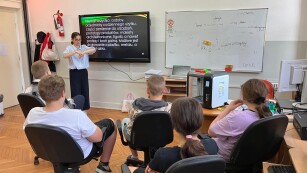 w sali informatycznej nauczycielka stoi przed monitorem multimedialnym, na którym pokazywana jest prezentacja. Uczniowie siedzą na krzesłach i patrzą na ekran