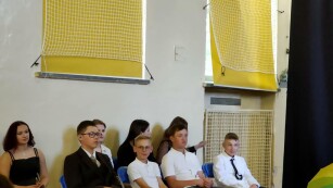 uczniowie w galowych strojach siedzą na krzesłach w sali gimnastycznej